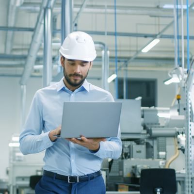 Chief Engineer in the Hard Hat Walks Through Light Modern Factory While Holding Laptop. Successful, Handsome Man in Modern Industrial Environment.